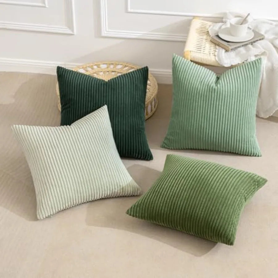 Four square ribbed throw pillows in different shades of green and light sage, arranged on a beige floor in a styled room setting with a woven stool and a small table in the background.